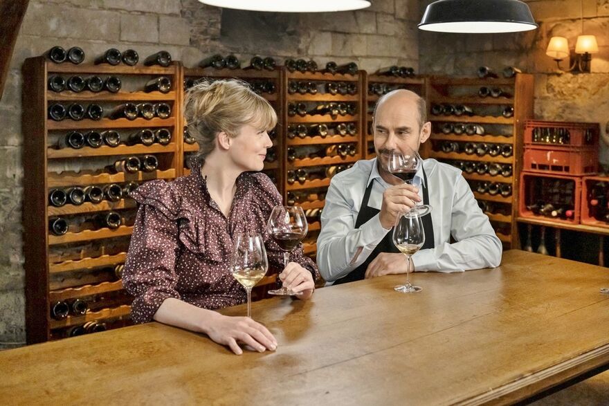 A man and a woman sit in a wine cellar, tasting glasses of wine and smiling at each other.