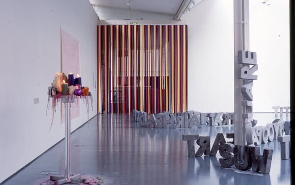 DCA Galleries during Fiona Banner's exhibition - a high table is covered in melting wax candles, large words written in grey concrete sit on the floor, and a vertical banner of colourful string hangs from the wall.