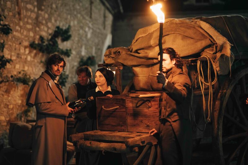 Three people dressed in clothes from the 1800s stand next to a cart in a dark street. One person is holding a large stick to light the way.