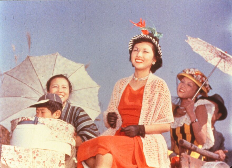 A Japanese lady in a beautiful red dress and fancy hat smiles as she is welcomed home on a sunny day