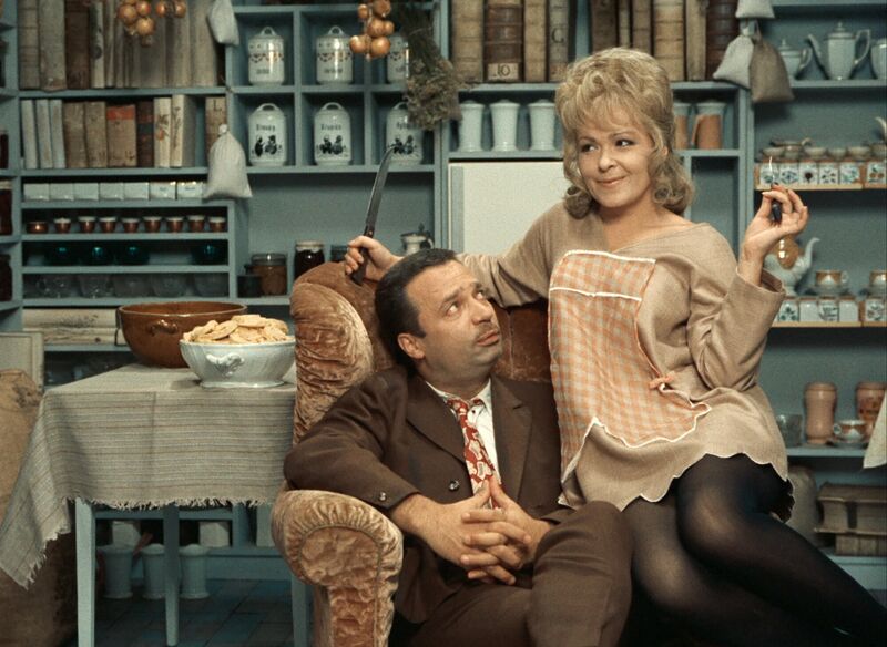 In a 1960s-style kitchen, a woman sits on a man's knee. The man is sitting on an arm chair. The woman is wearing an apron and smiling.