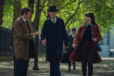 Two men and a woman, wearing clothing from the nineteen sixties, walk down a tree-lined street.