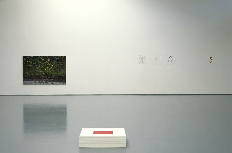 A white square rests on a grey floor. A green, abstract canvas hangs on a white wall, and small drawings are also pinned to the wall.