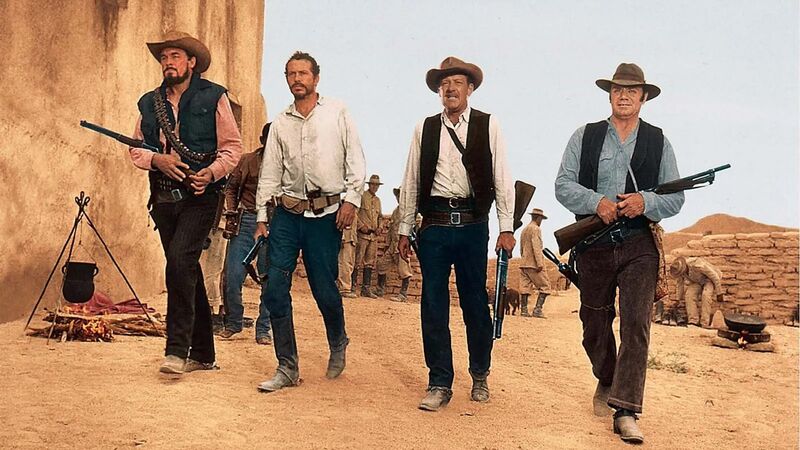 Four cowboys holding guns walk through a sandy town.