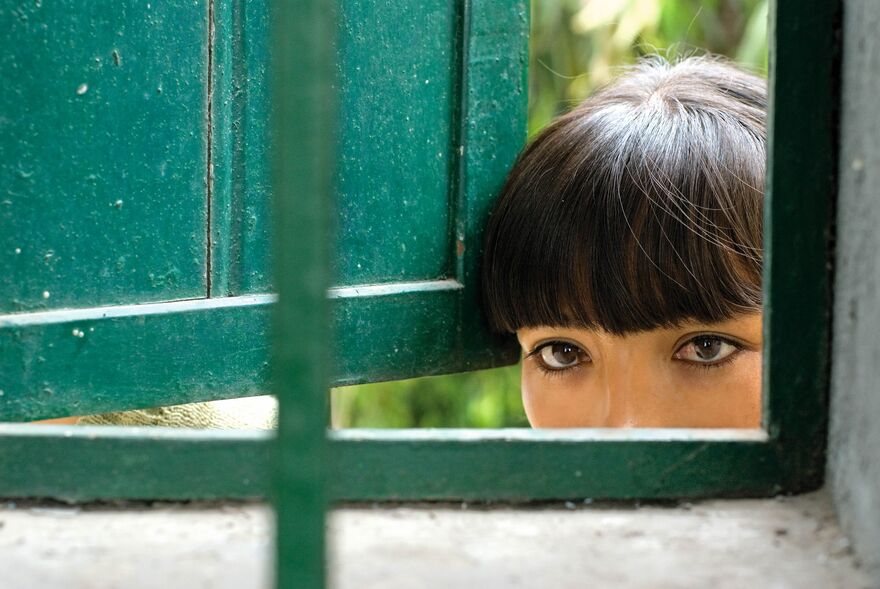 A young woman with dark hair peeks through a window. She can be seen from the nose up.