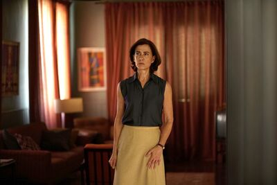 A serious woman stands in a living room, wearing a yellow skirt.