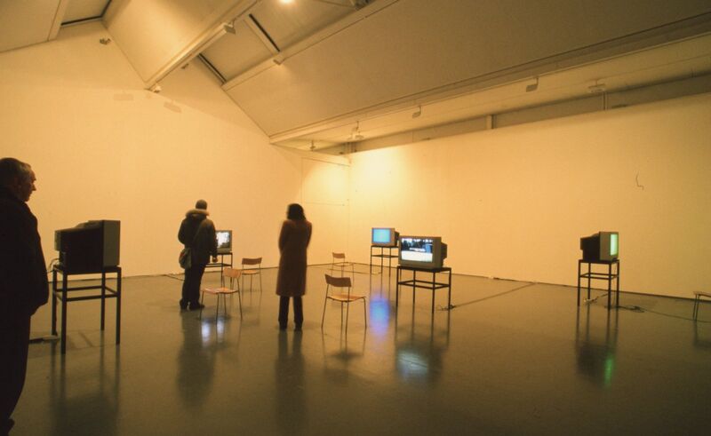 People stand in a white gallery space filled with televisions and wooden chairs.