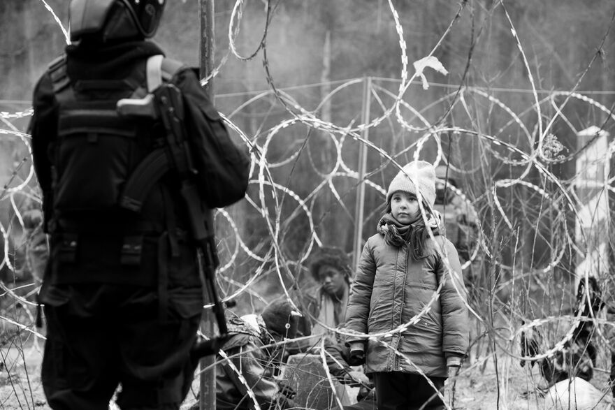 A girl in winter clothing stands behind razor wire, looking at a soldier