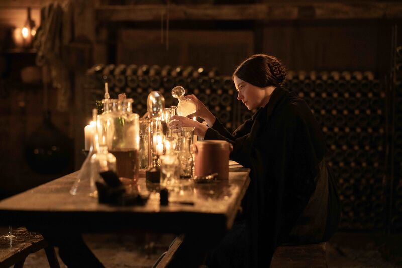 A woman sits at a desk by candlelight, pouring a liquid into various receptacles in a scientific manner.