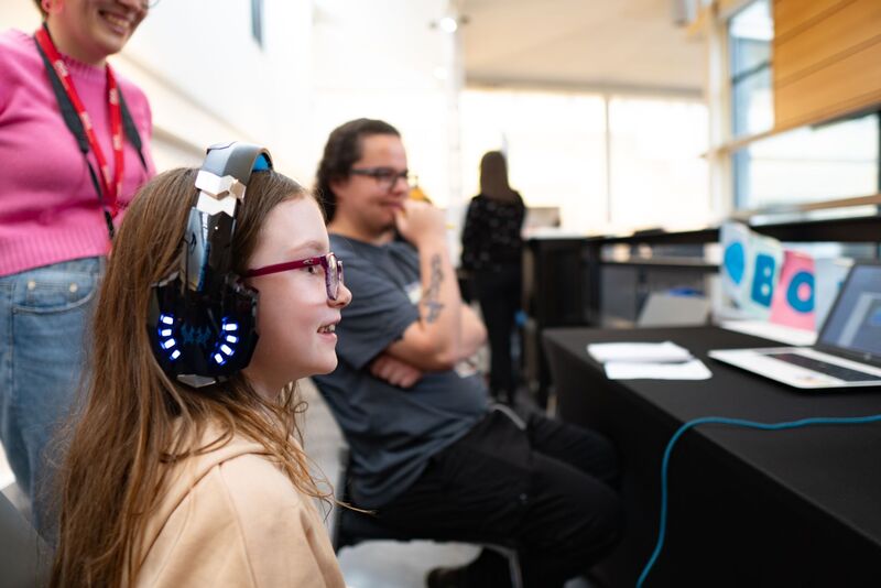 A young child plays a game wearing large headphones at Drop in and Play while people watch.