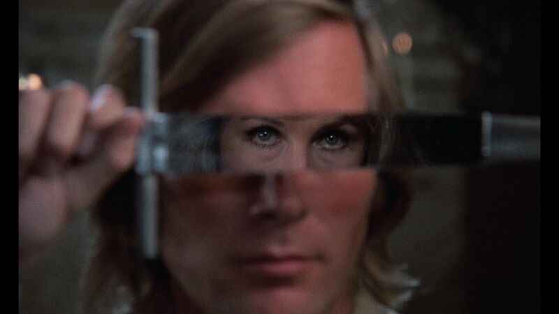 A man holds a dagger in front of his face. In the reflection, we can see the eyes of a woman.