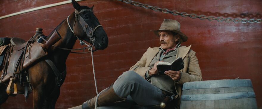 A man dressed in cowboy-esque clothes sits next to a barrel, holding a book. There is a horse next to him.