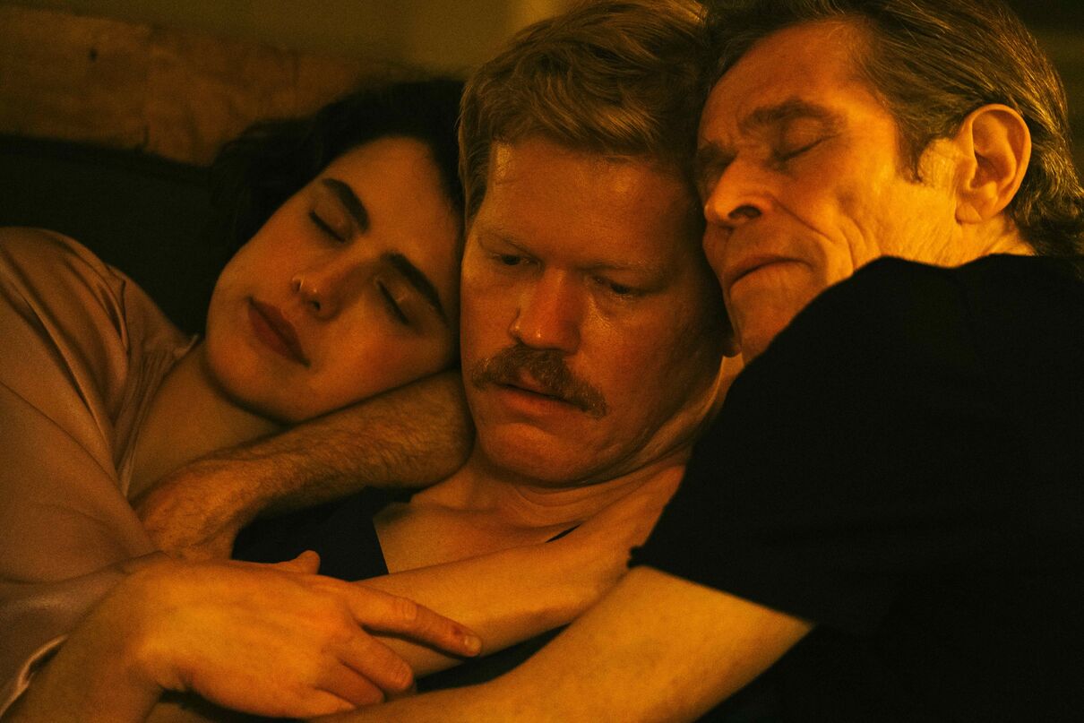 Three people cuddle in an orange-lit room. The person in the middle looks worried.