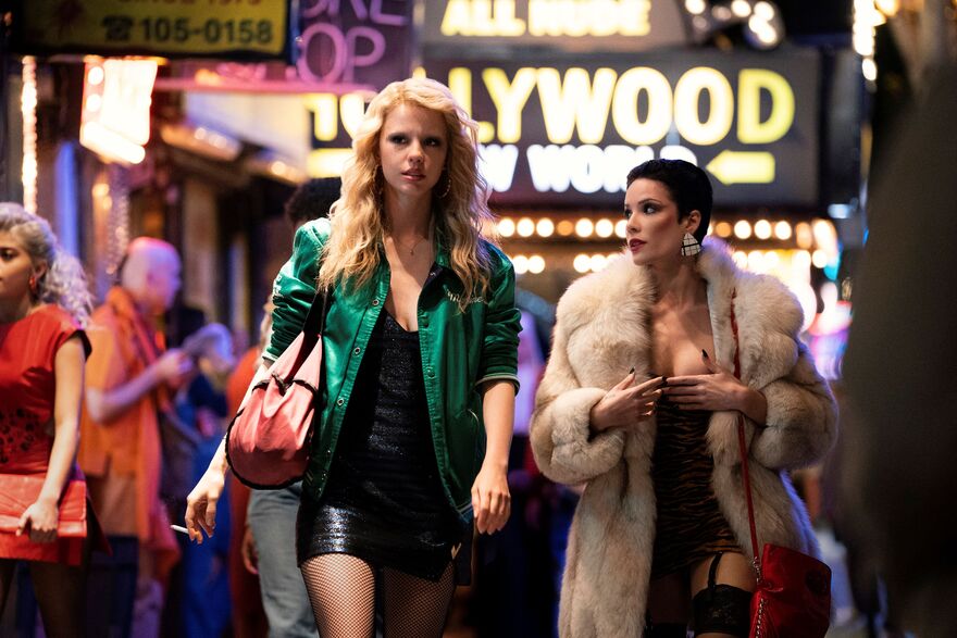 A tall blonde woman walks down a brightly lit street, next to a shorter woman wearing a fur coat.