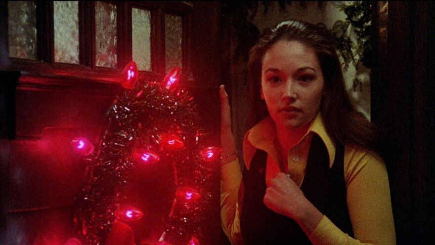 A woman with long brown hair stands next to a door with a wreath made of black wire and red glowing Christmas lights.