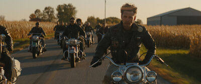 A group of people in leathers ride motorcycles down a rural, sunny road.