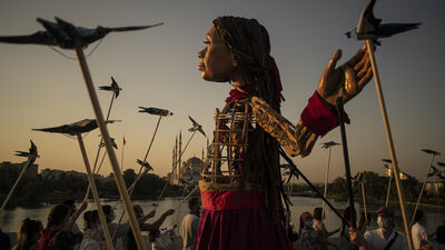 A huge puppet girl walks at sunset, surrounded by puppet birds