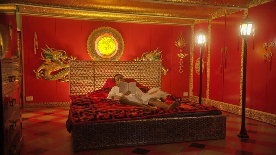A person lies on a red bed in a room decorated with red walls and gold dragons, reading a book.
